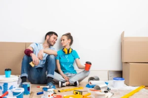 Couple souriant entouré de matériel de chantier et de cartons, illustrant comment payer moins cher la rénovation de son appartement.